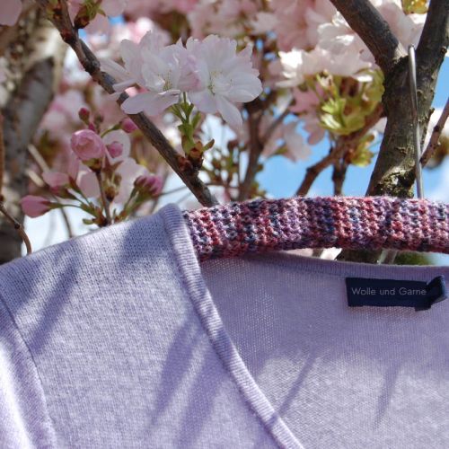 Fliederfarbene Strickjacke auf umhäkeltem Kleiderbügel hängt im blühenden Kirschbaum